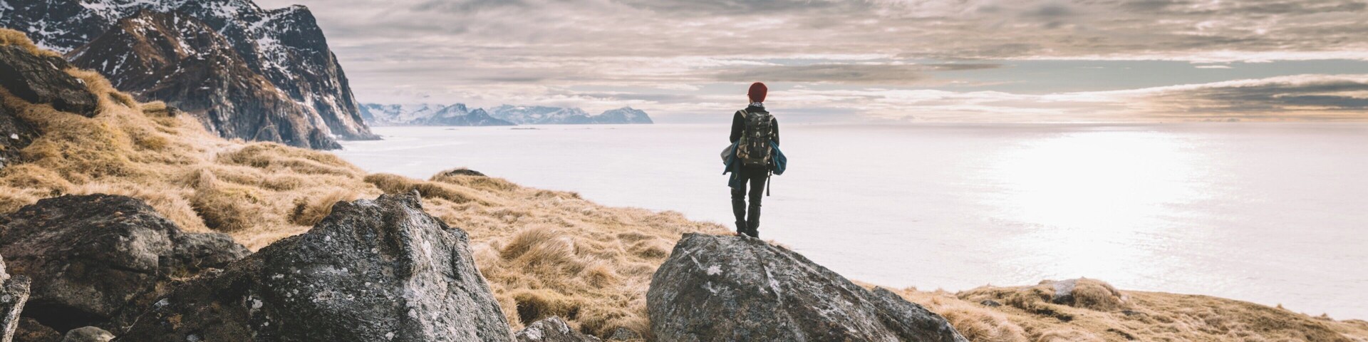 A person standing on a rocky outcrop overlooking a calm body of water with mountains in the distance.