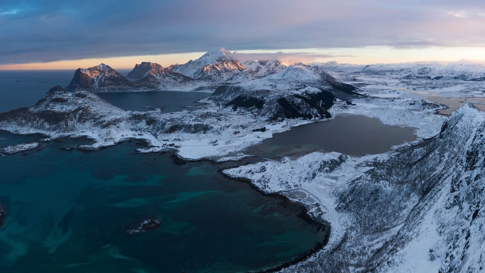 A snow-covered mountainous landscape with a fjord and clear blue waters.