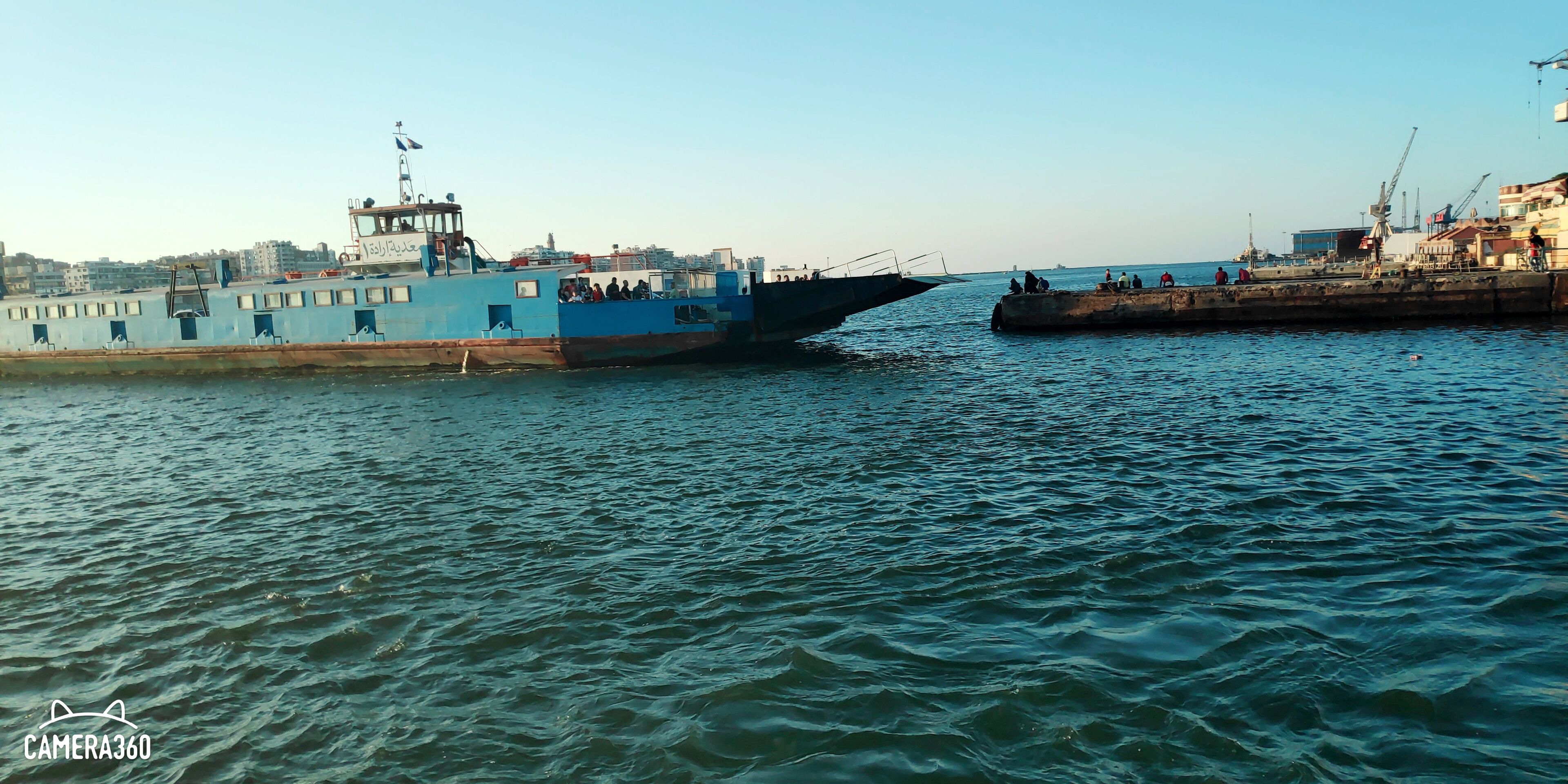 A large blue ship docked at a port with a pier and buildings in the background.