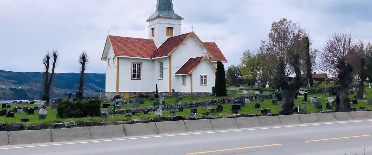 A white church with a red roof and a tall steeple, surrounded by a cemetery with gravestones.
