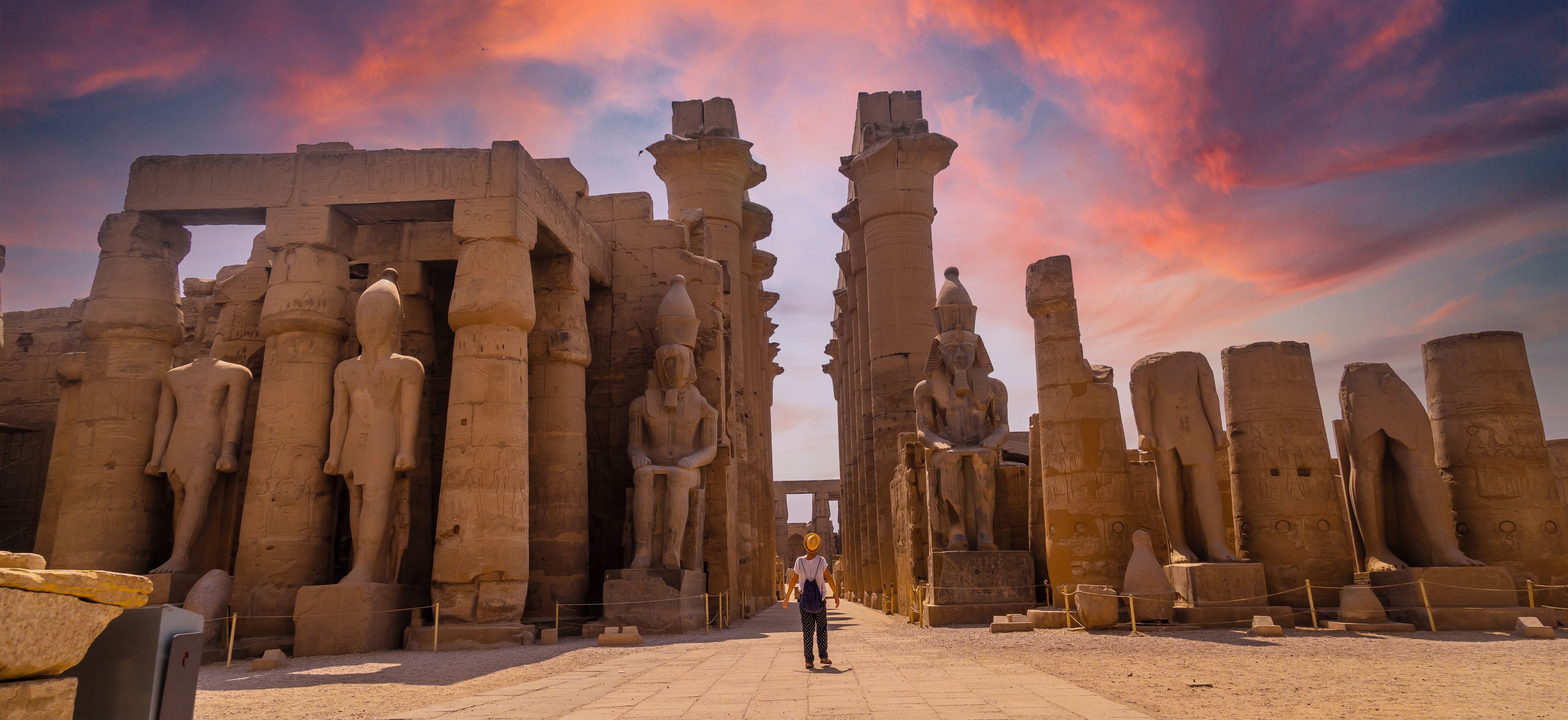 Ancient stone ruins with columns and statues, a person walking through the site, and a sunset sky.