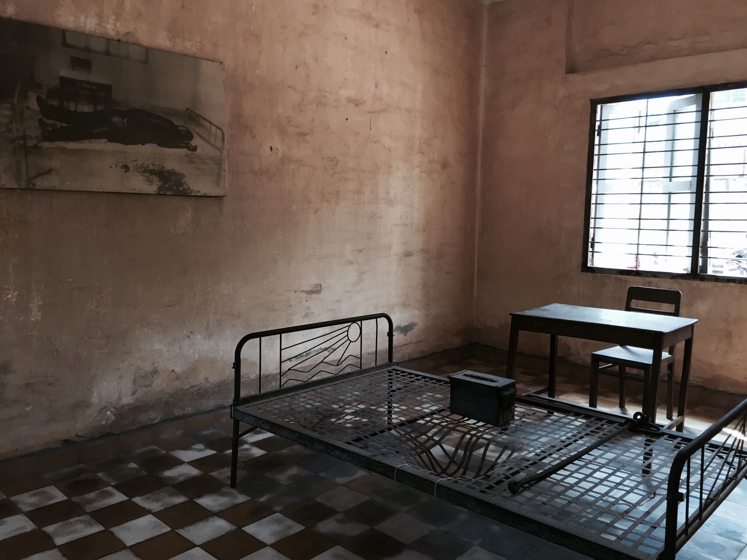 A room with a metal bed frame, a table, and a chair. There is a window with bars and a painting on the wall.