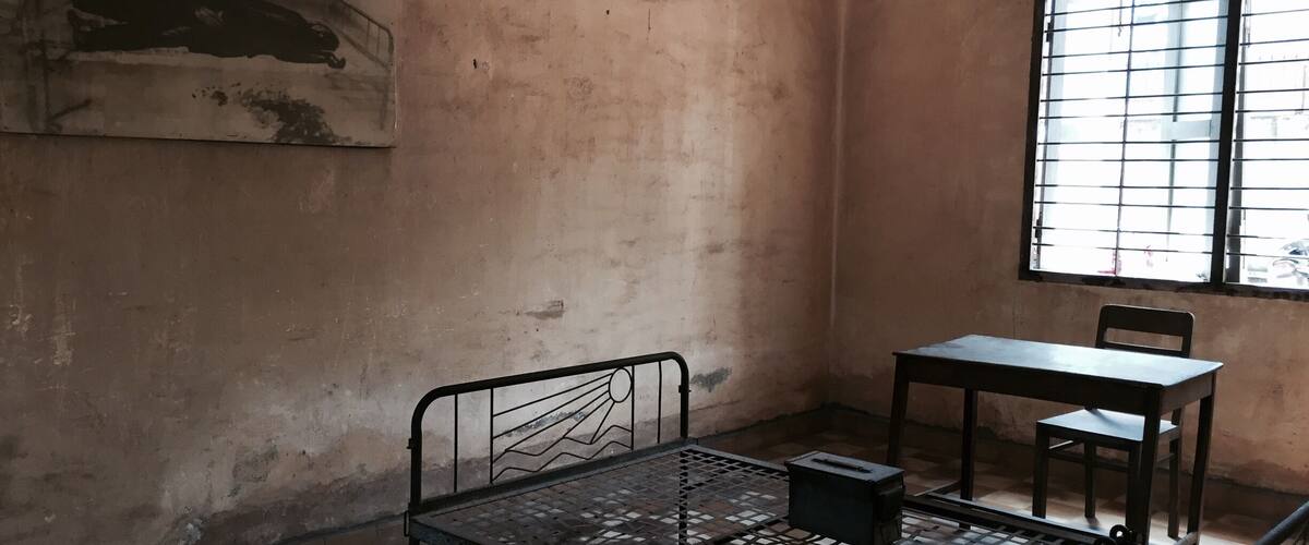 A room with a metal bed frame, a table, and a chair. There is a window with bars and a painting on the wall.