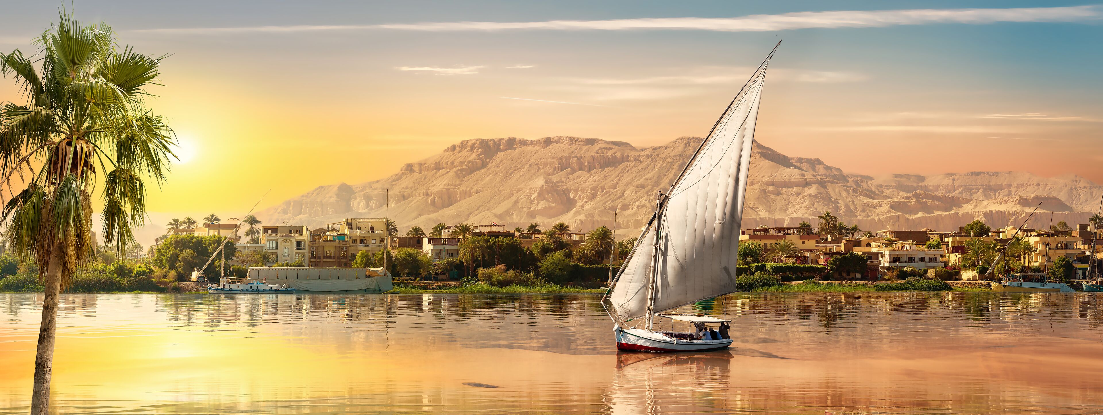 A sailboat on calm waters with a backdrop of a mountainous landscape and a town with traditional buildings.