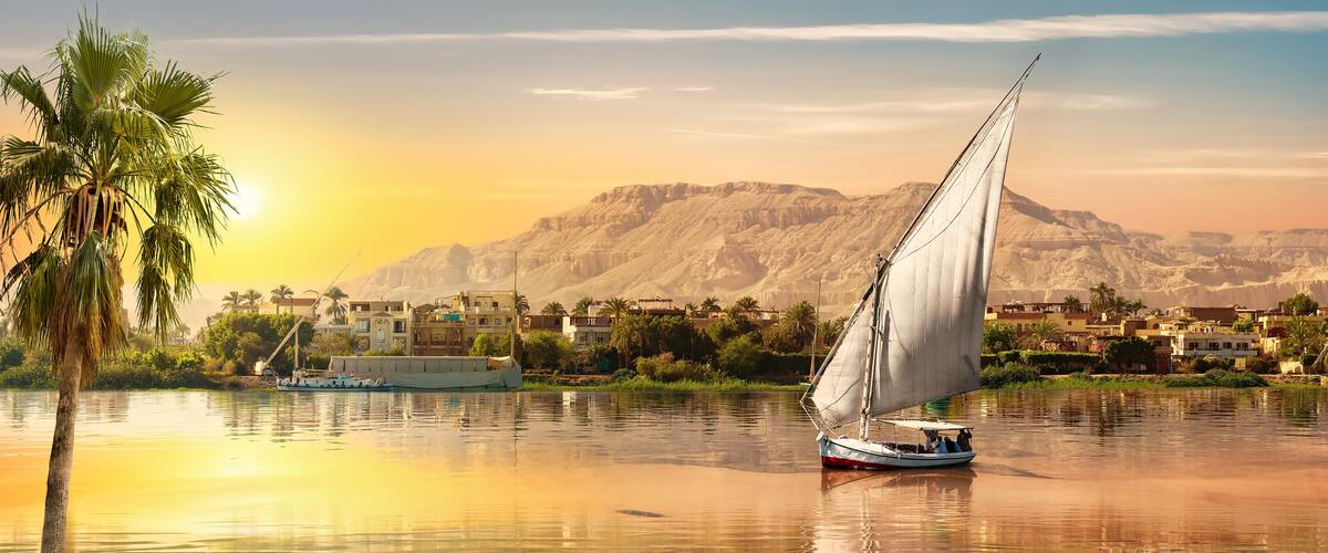 A sailboat on calm waters with a backdrop of a mountainous landscape and a town with traditional buildings.