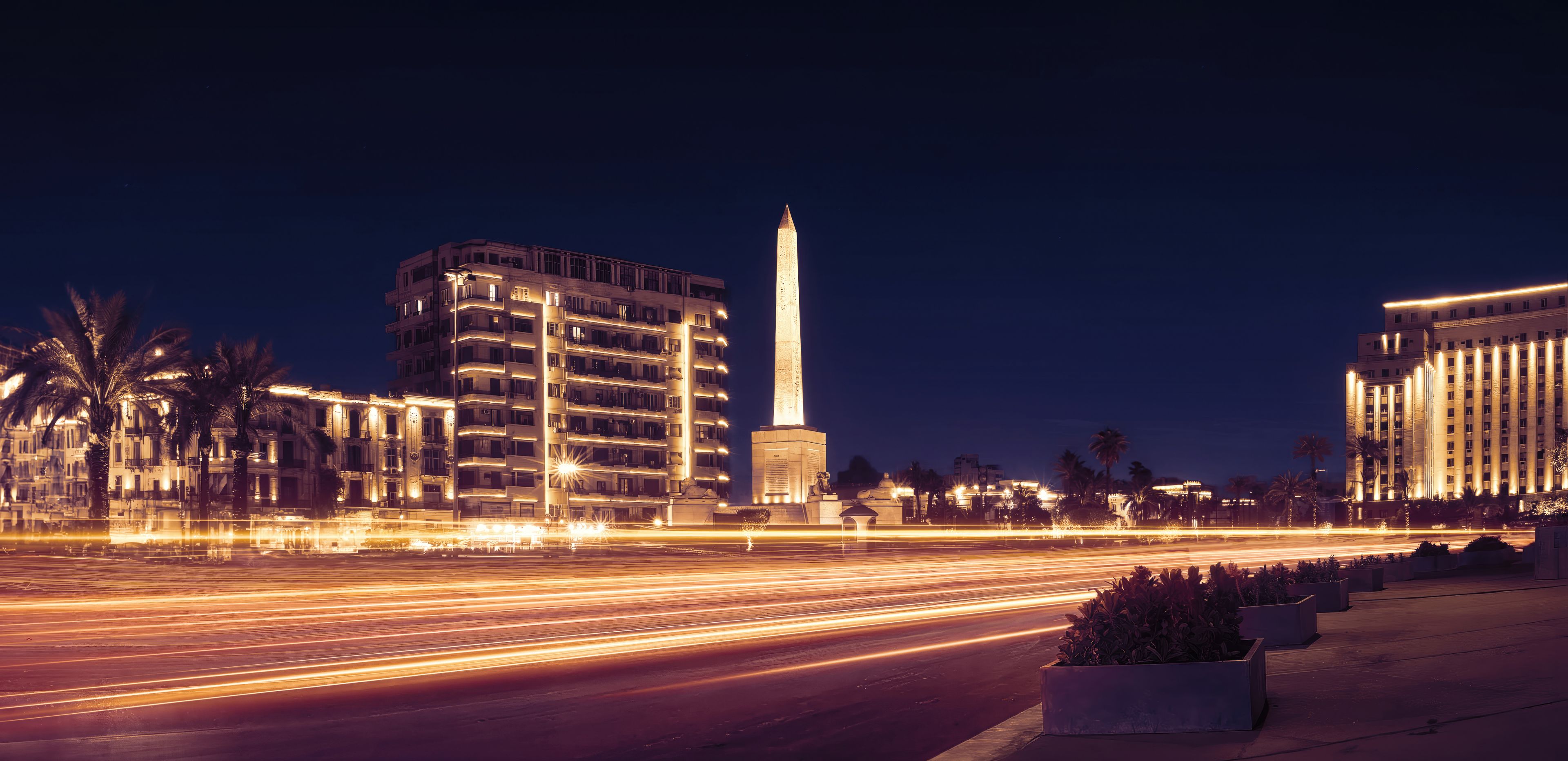 Un paisaje urbano nocturno con edificios iluminados, un monumento alto y estelas de luz dejadas por vehículos en movimiento.