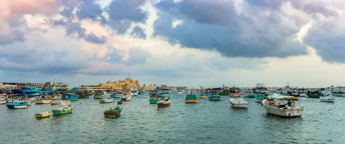 ALEXANDRIA, EGYPT - NOVEMBER 20, 2016: Cityscape with egyptian fisher boats, vessels and ships in the seaport at sunrise. Panoramic view with Qaitbay Citadel in a harbour of Alexandria.