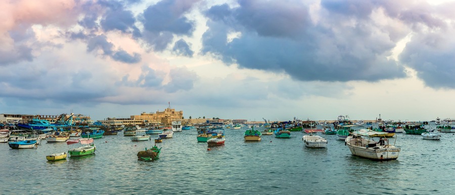 ALEXANDRIA, EGYPT - NOVEMBER 20, 2016: Cityscape with egyptian fisher boats, vessels and ships in the seaport at sunrise. Panoramic view with Qaitbay Citadel in a harbour of Alexandria.
