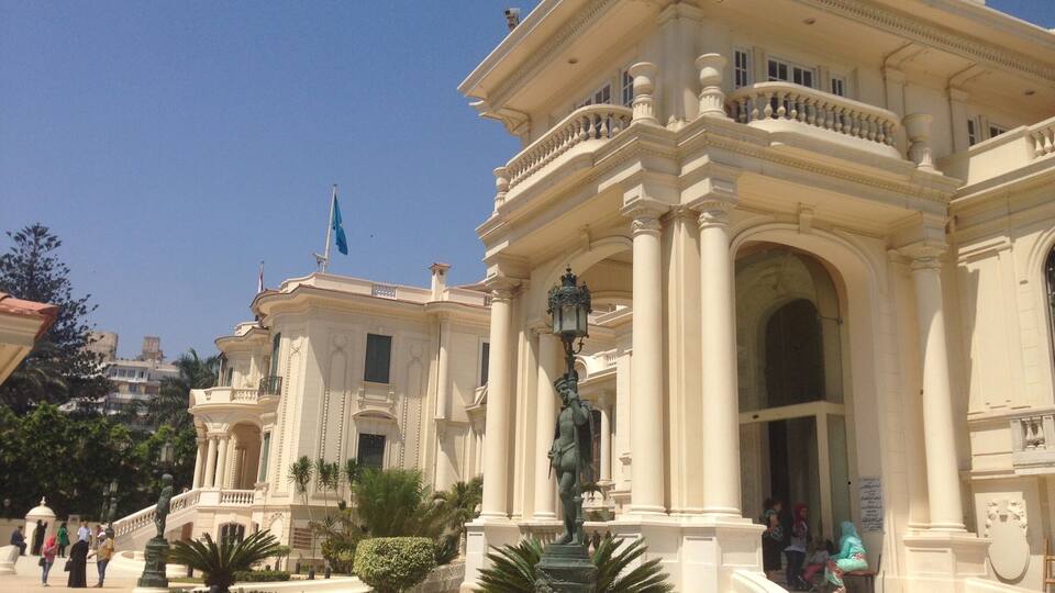 Un edificio imponente con arquitectura clásica, una estatua y una fuente en una plaza ajardinada.