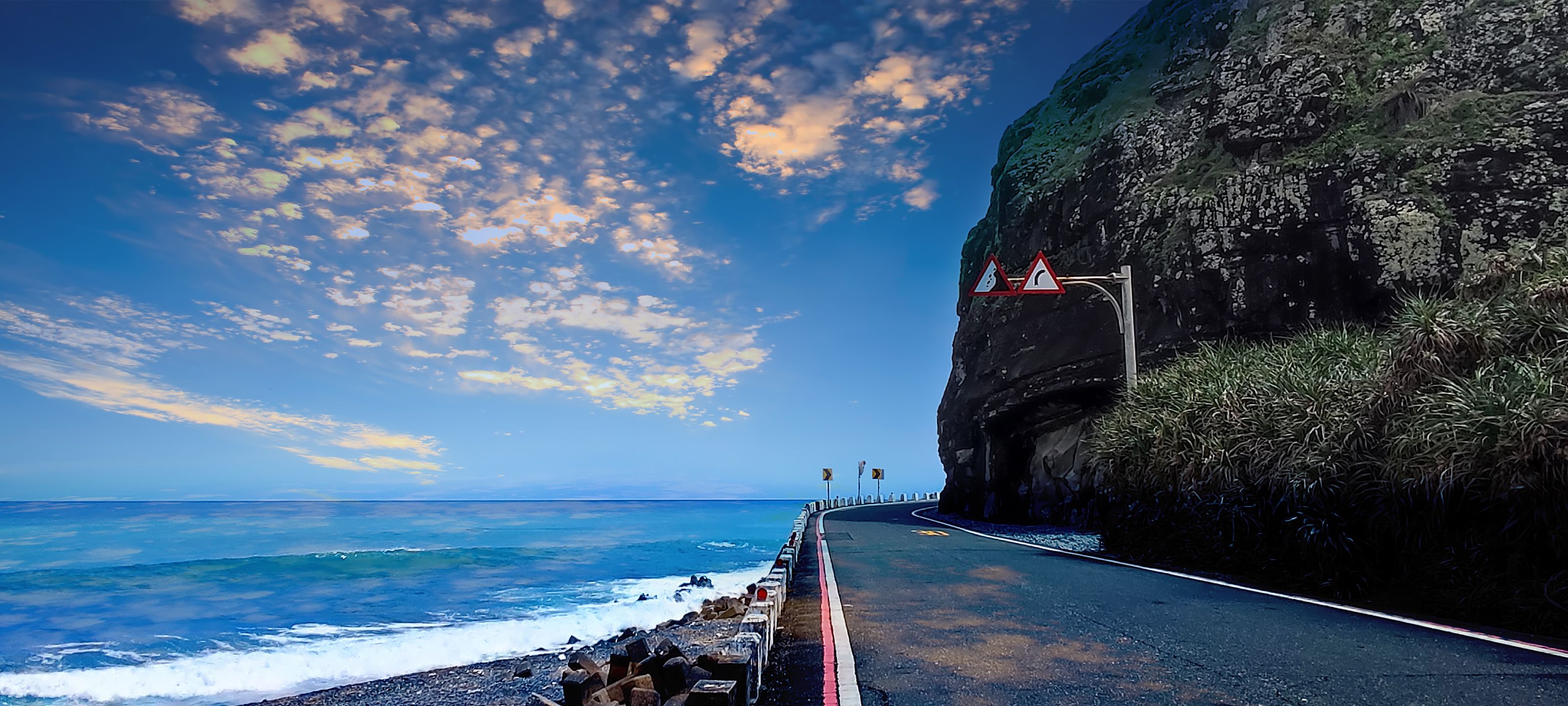 A coastal road with a cliffside, ocean waves, and a clear sky.