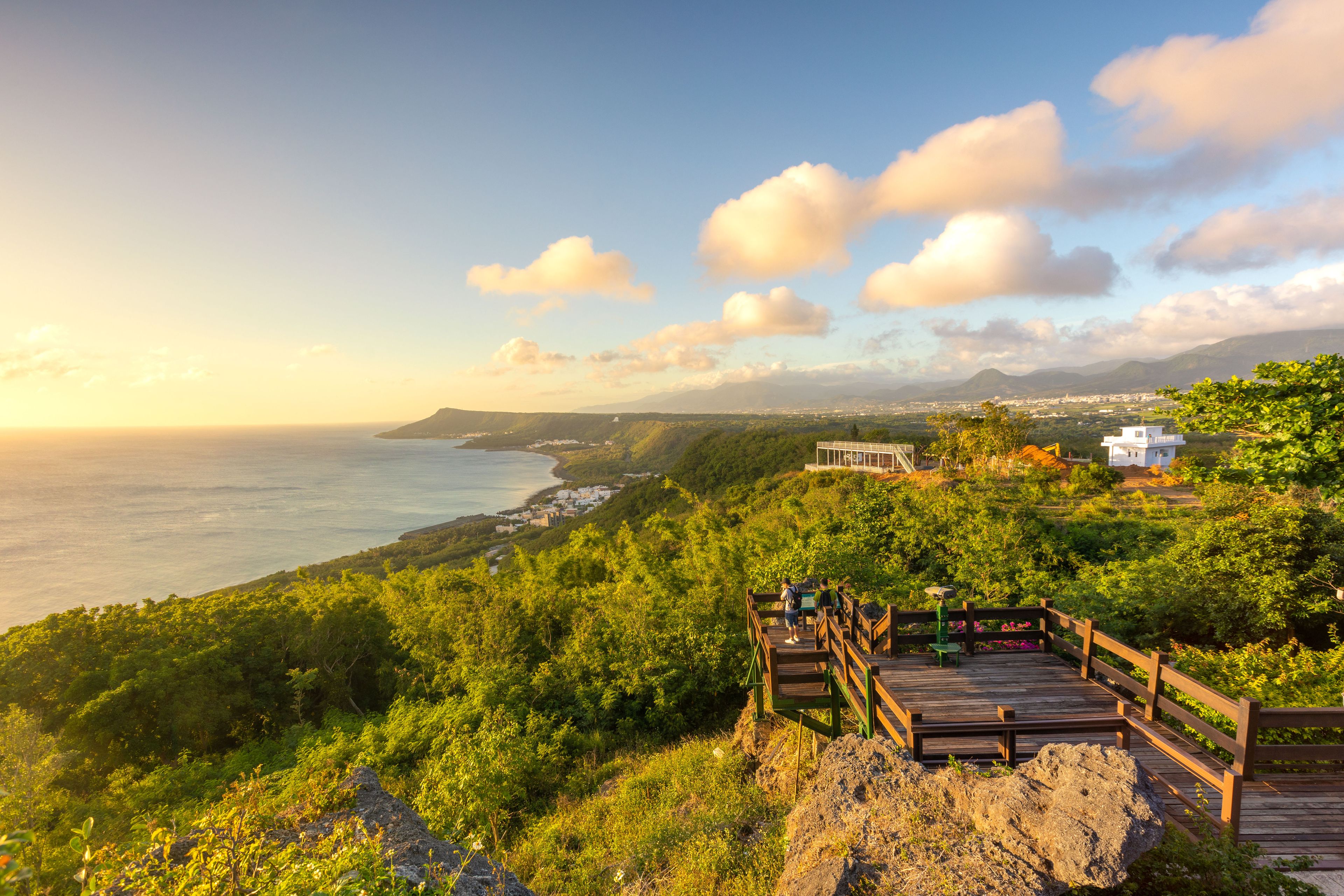 A coastal landscape with a wooden viewing platform overlooking the sea, lush greenery, and distant mountains.