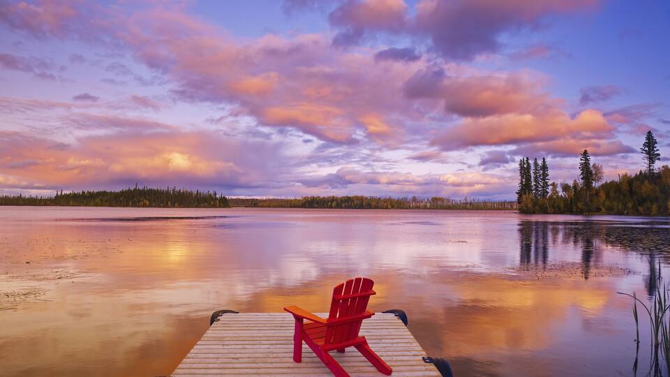 A wooden dock with a red Adirondack chair facing a calm lake at sunset.