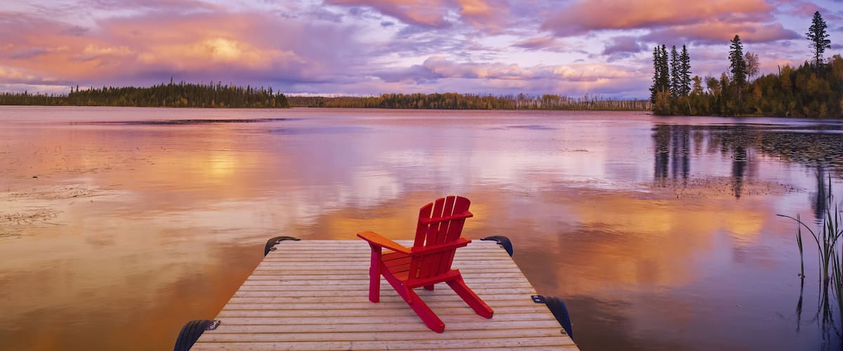 A wooden dock with a red Adirondack chair facing a calm lake at sunset.