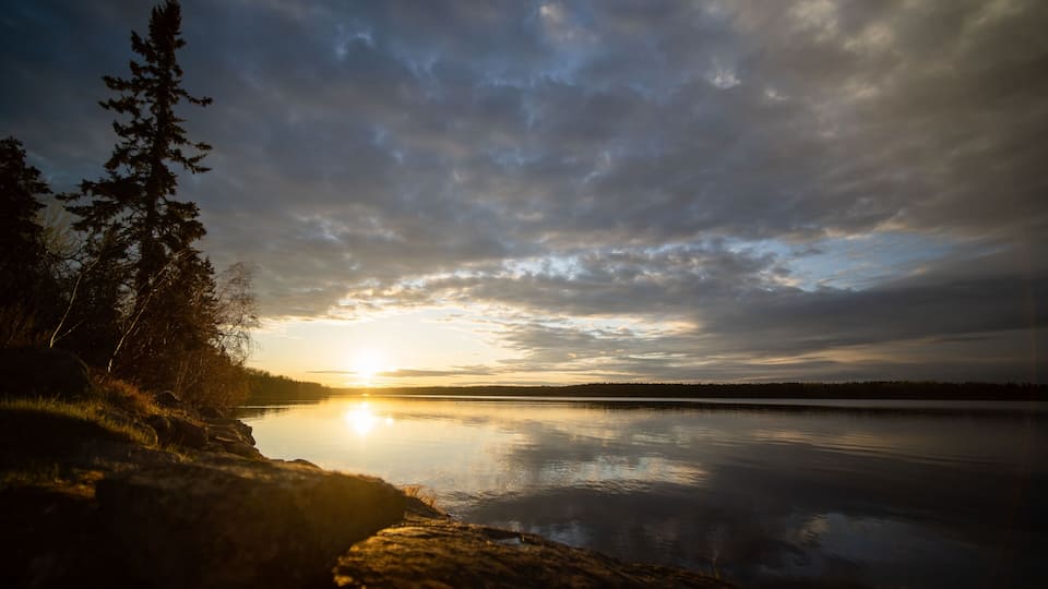 A sunset over a calm lake with trees on the left and a cloudy sky.