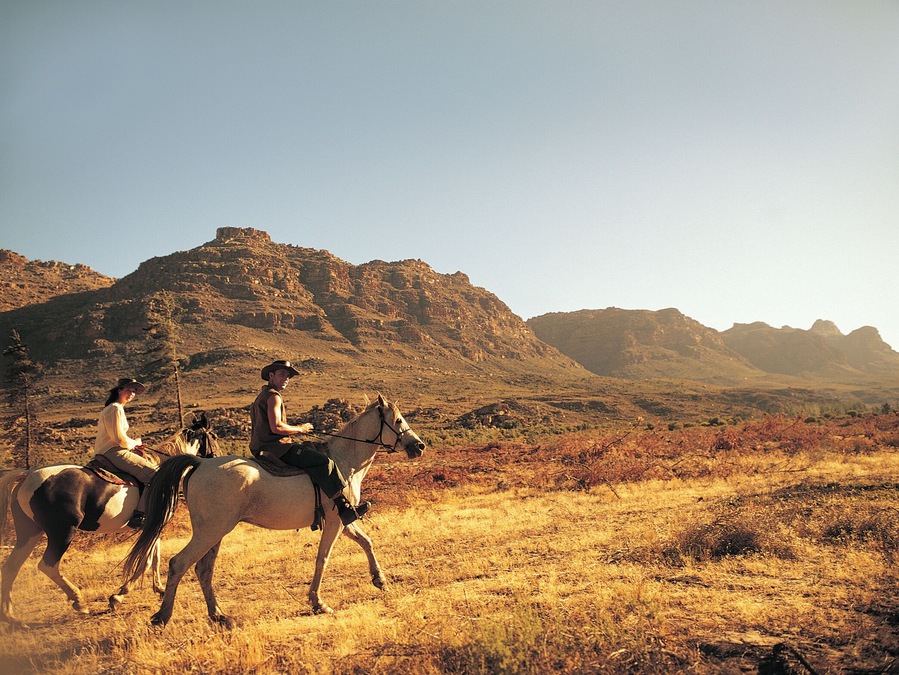 Couple Riding Horses Through Scrubland With Rugged Hills in the Background in Africa