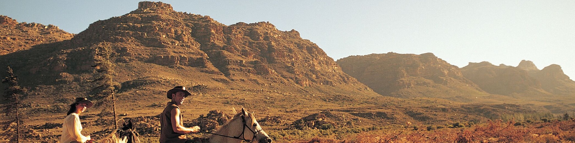 Couple Riding Horses Through Scrubland With Rugged Hills in the Background in Africa