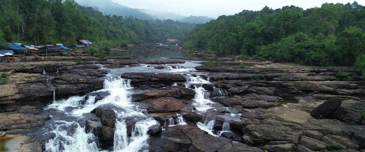 A river with multiple waterfalls flowing over rocks, surrounded by dense forest and a few structures on the left.