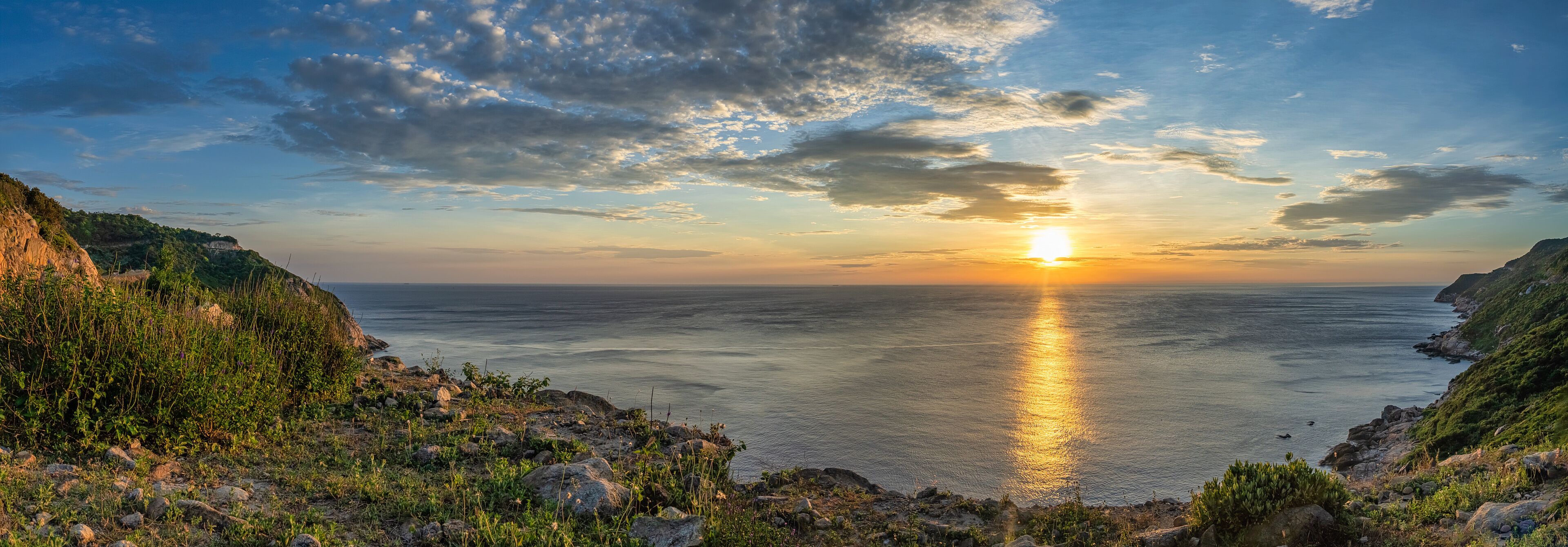 A coastal sunset with the sun reflecting on the water, rocky cliffs, and sparse vegetation.