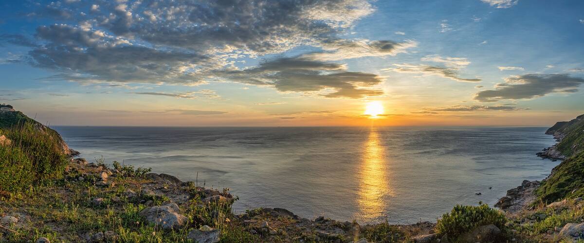A coastal sunset with the sun reflecting on the water, rocky cliffs, and sparse vegetation.