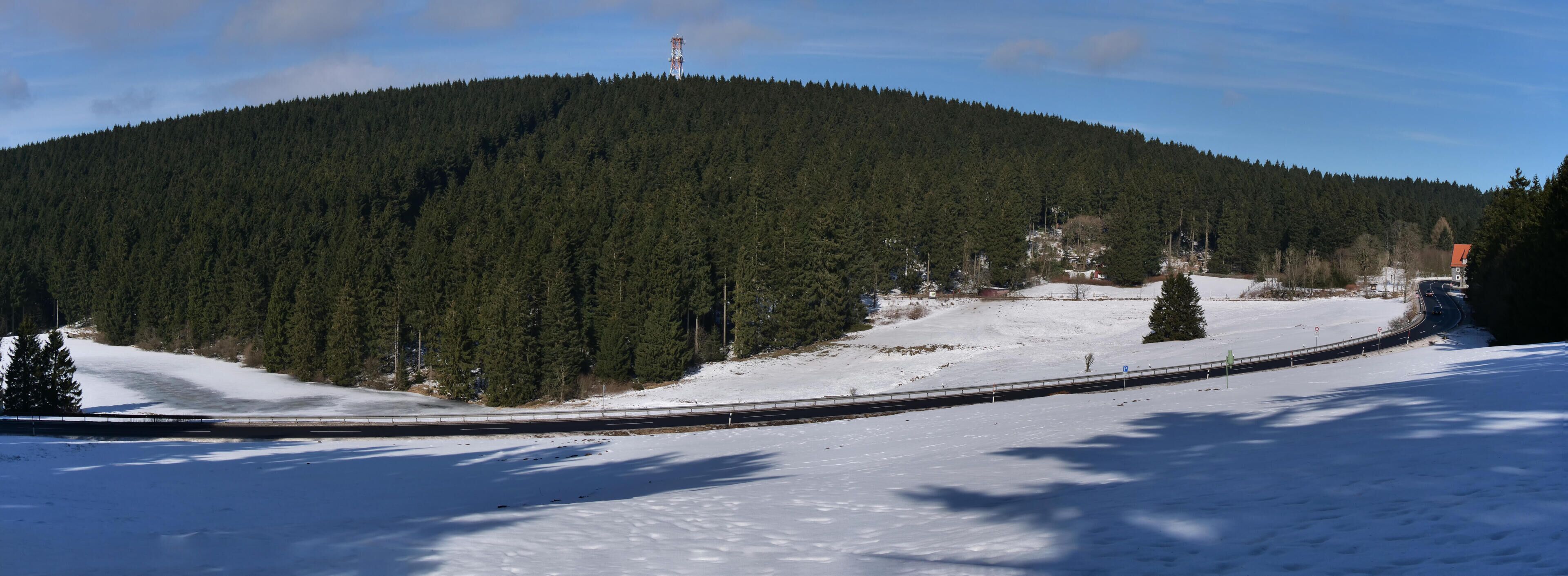 A snow-covered landscape with a forested hill, a road curving through the scene, and a small building with a red roof.