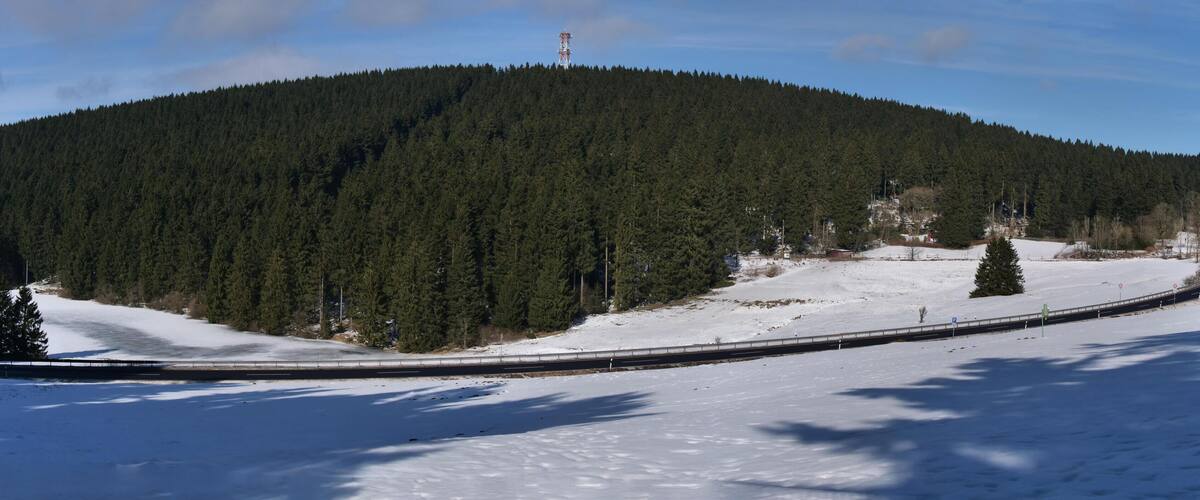 A snow-covered landscape with a forested hill, a road curving through the scene, and a small building with a red roof.