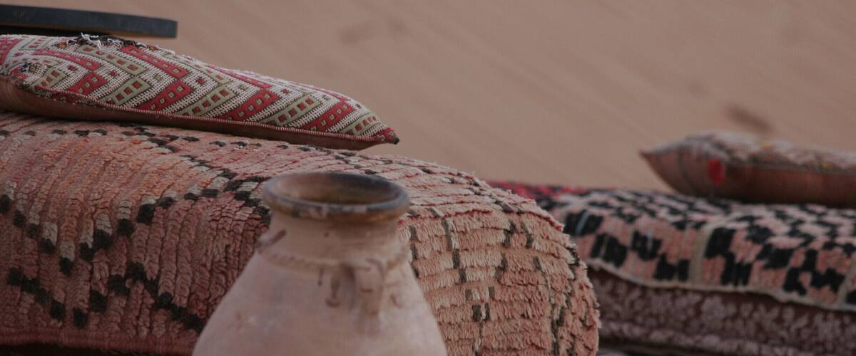 A traditional woven rug, a clay pot, and a patterned cushion on a sandy surface.