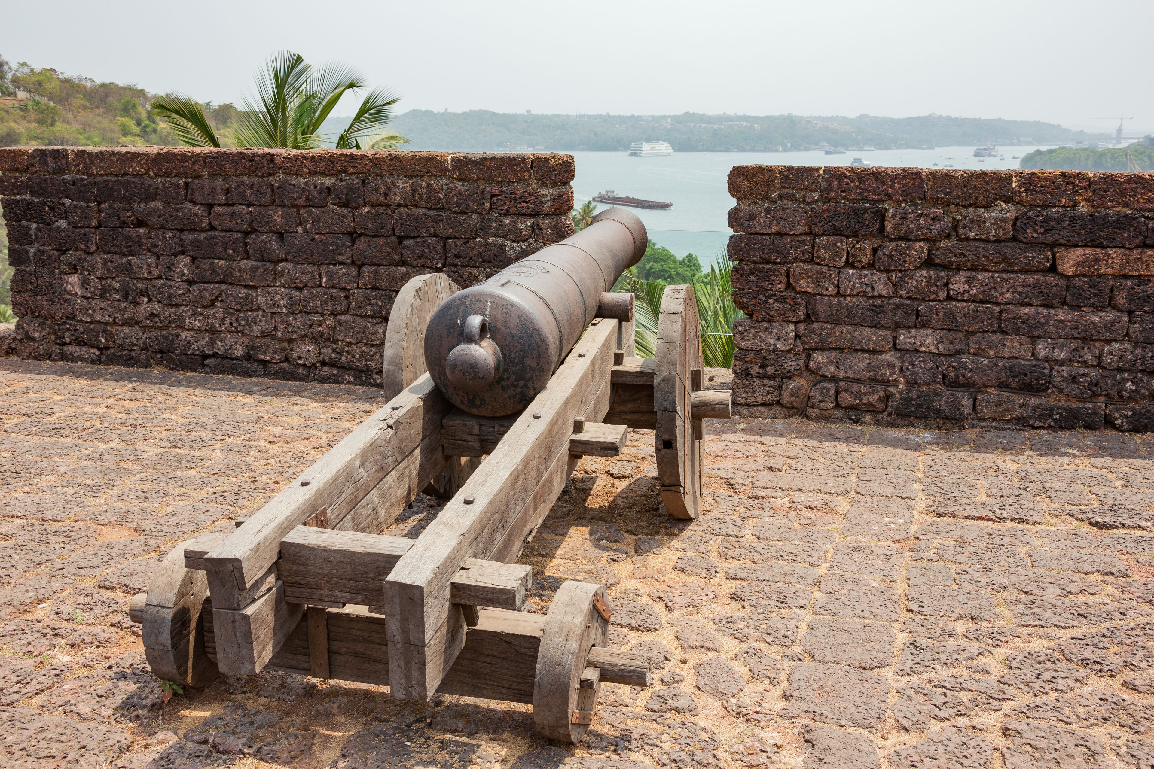 Um canhão antigo montado em um suporte de madeira, posicionado entre duas paredes de tijolo com vista para um corpo d'água com barcos e uma linha costeira distante.