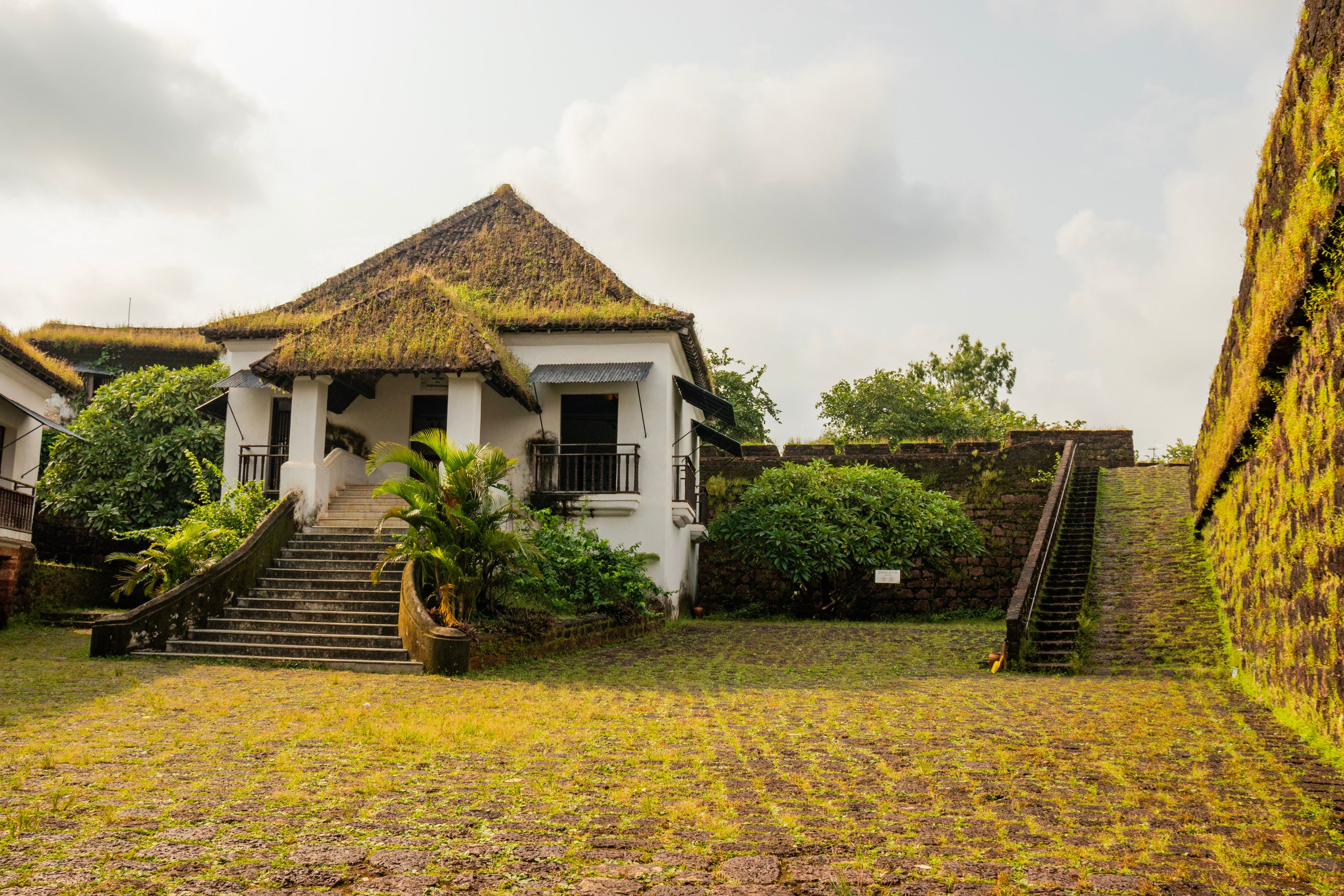 Um edifício de estilo colonial branco, com telhado de palha, rodeado por vegetação exuberante e uma escadaria que leva até ele.