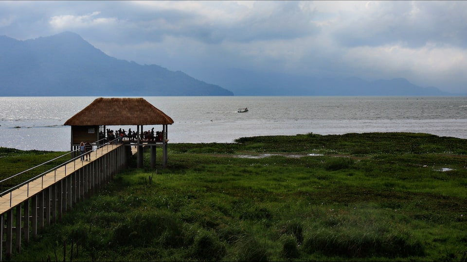 A wooden viewing platform with a thatched roof overlooking a lake, with mountains in the background.