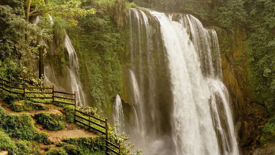 A large waterfall surrounded by lush greenery and a wooden walkway.