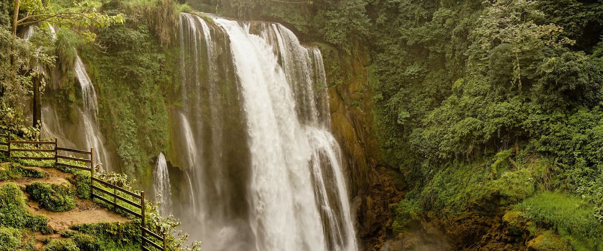 A large waterfall surrounded by lush greenery and a wooden walkway.