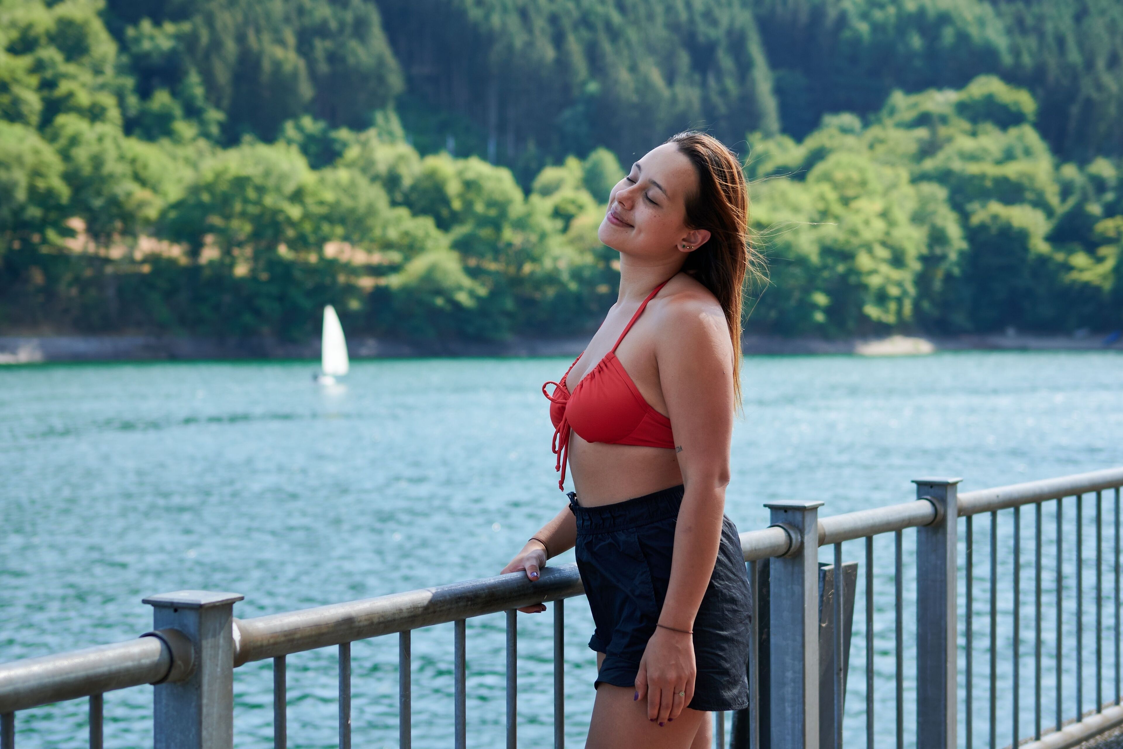 A person standing by a railing overlooking a lake with a forested shoreline and a sailboat in the distance.