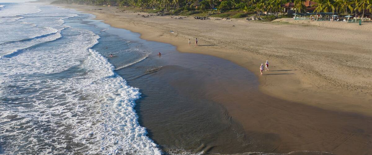 A beach with waves, palm trees, and mountains in the background.
