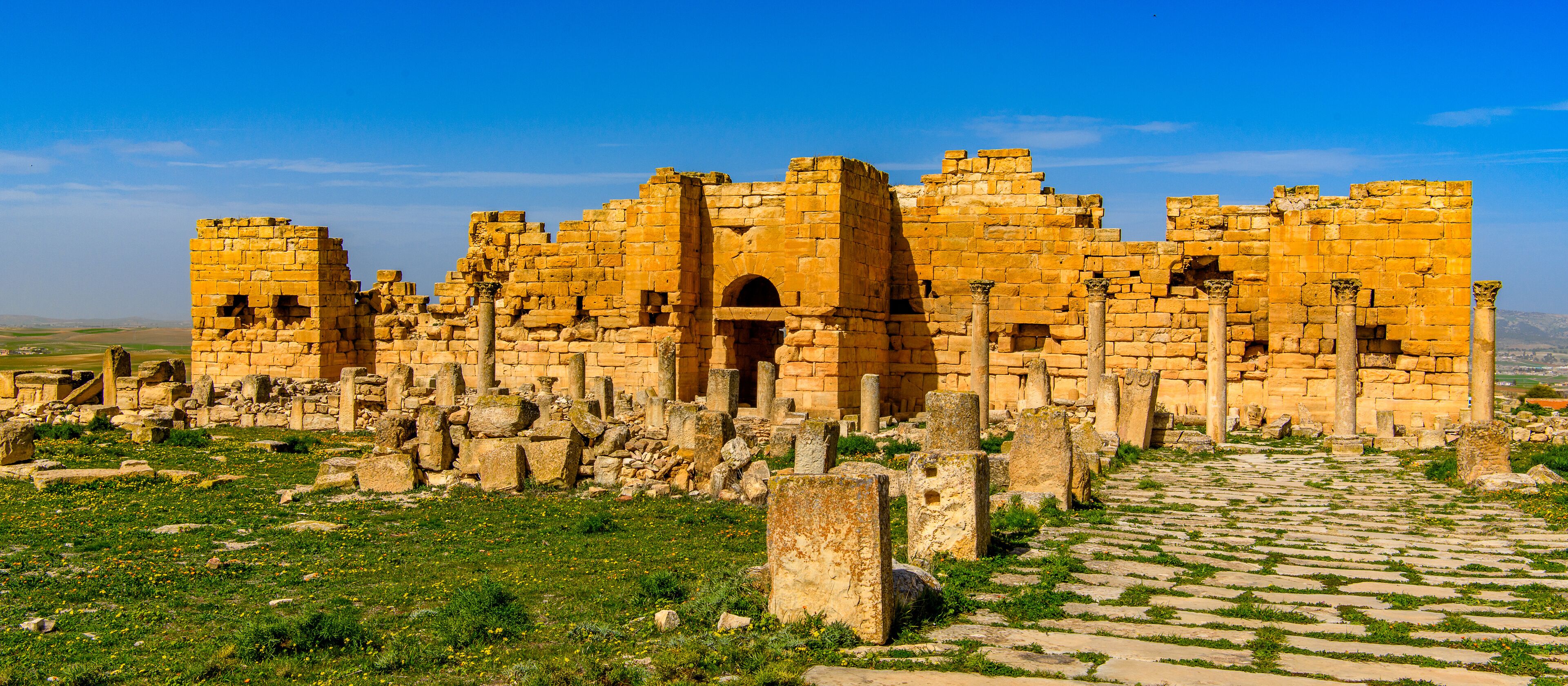 Ancient ruins with stone walls and columns, a cobblestone path, and a clear blue sky.