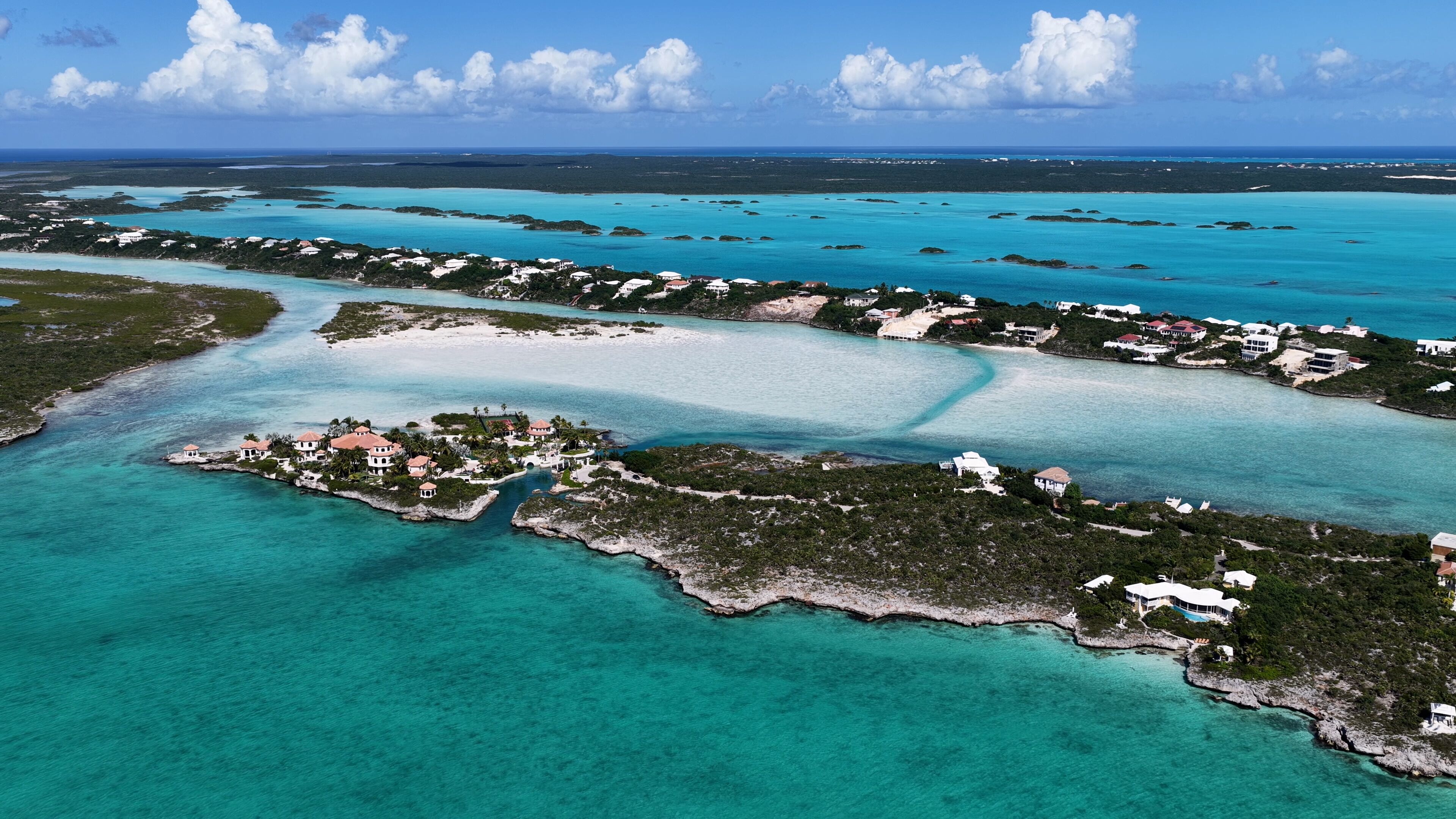 An aerial view of a coastal area with clear turquoise waters, a small island with buildings, and a distant shoreline.