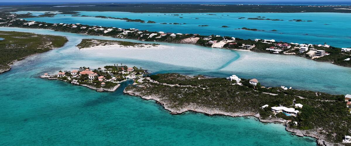 An aerial view of a coastal area with clear turquoise waters, a small island with buildings, and a distant shoreline.
