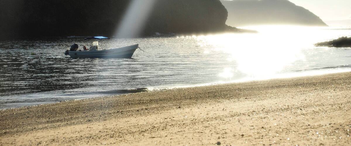 A boat on calm water near a sandy beach with a large rock formation in the background.
