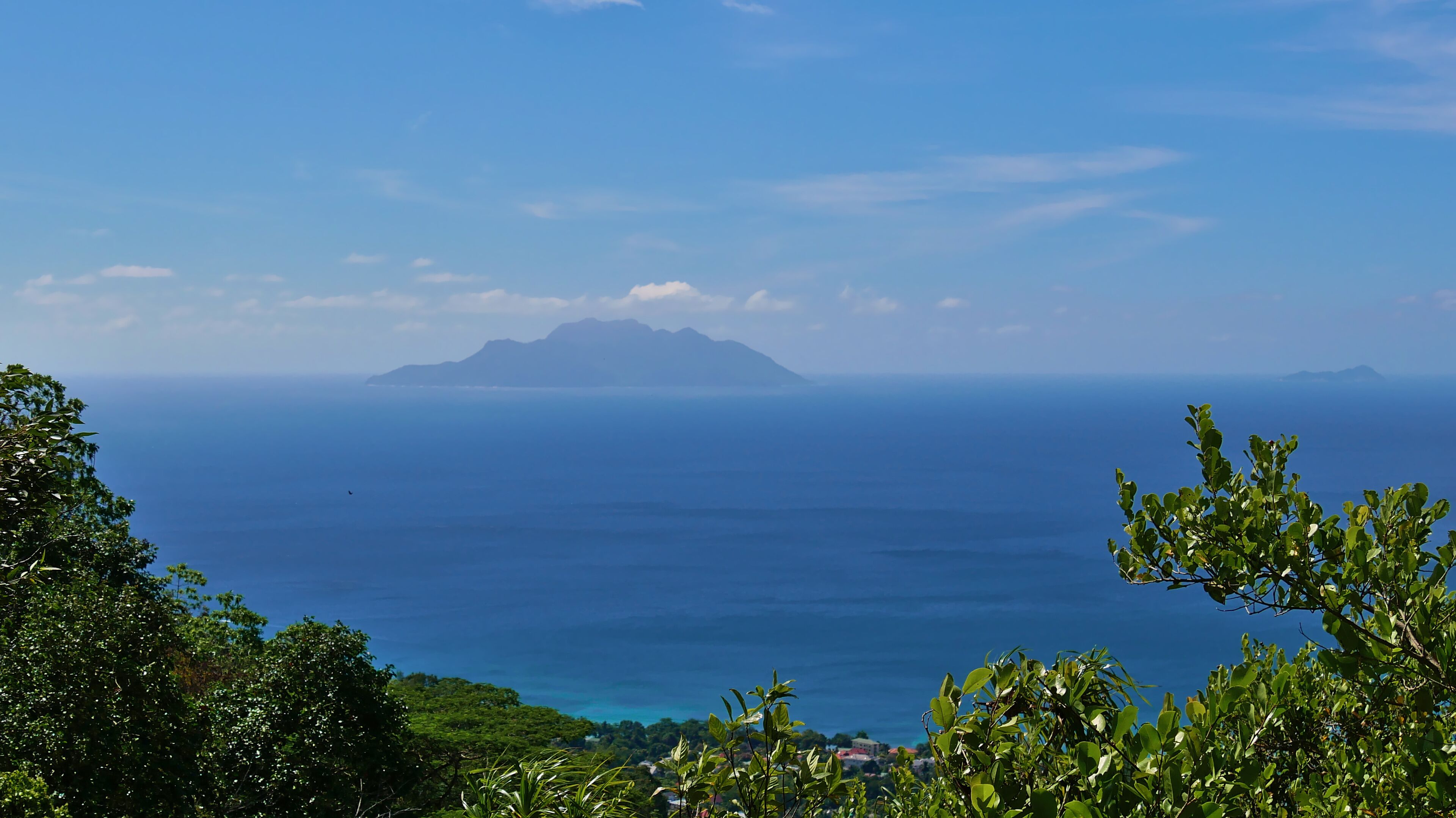 A coastal landscape with a mountain in the distance, clear blue sea, and lush greenery.