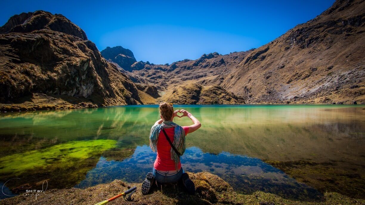 A person making a heart shape with their hands by a clear lake surrounded by mountains.