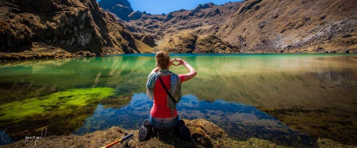 A person making a heart shape with their hands by a clear lake surrounded by mountains.