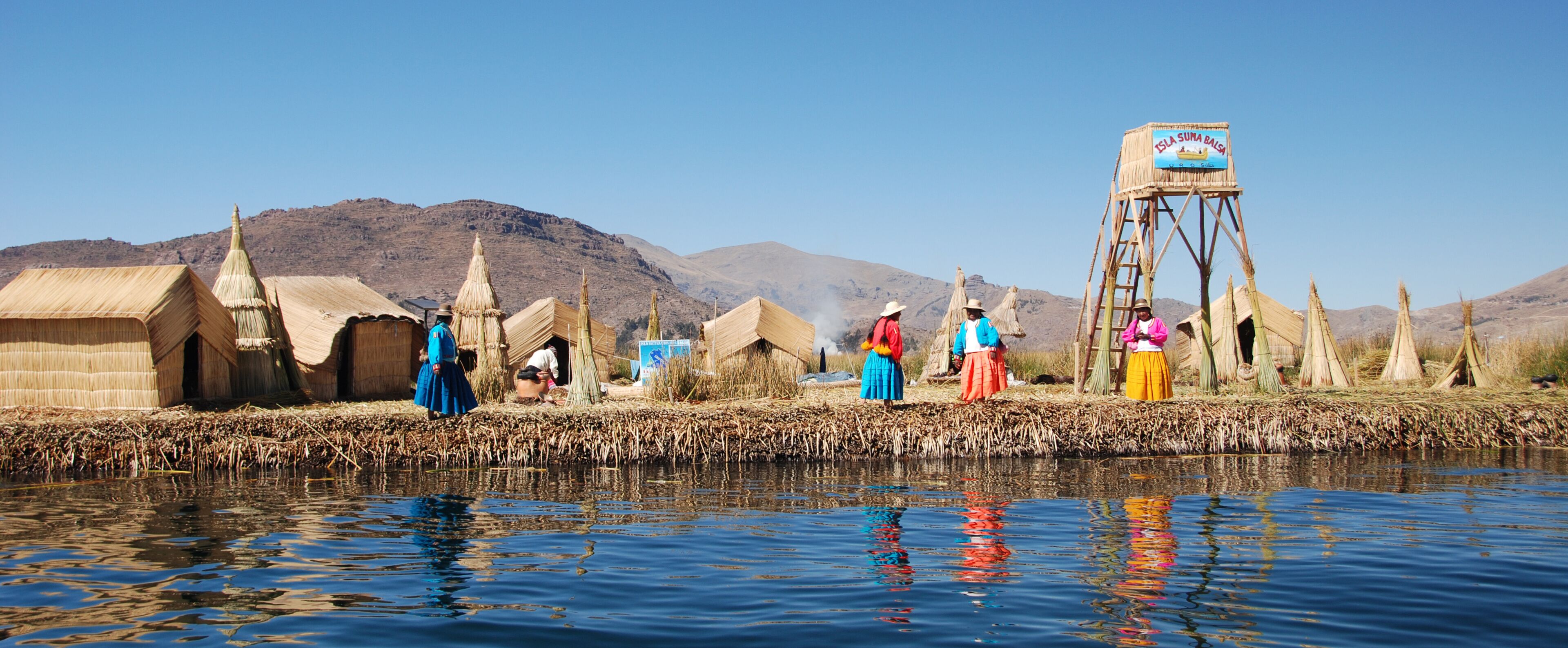 Traditional huts by a river, a water tower, and mountains in the background.