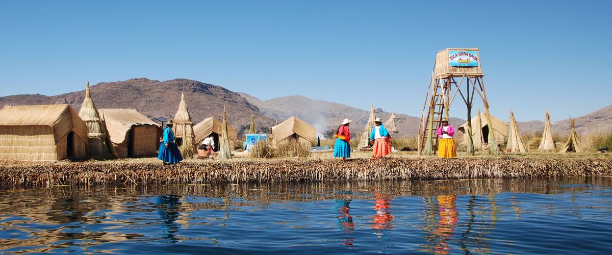 Traditional huts by a river, a water tower, and mountains in the background.