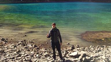 A person standing on a rocky shore with a clear turquoise lake and snow-capped mountains in the background.
