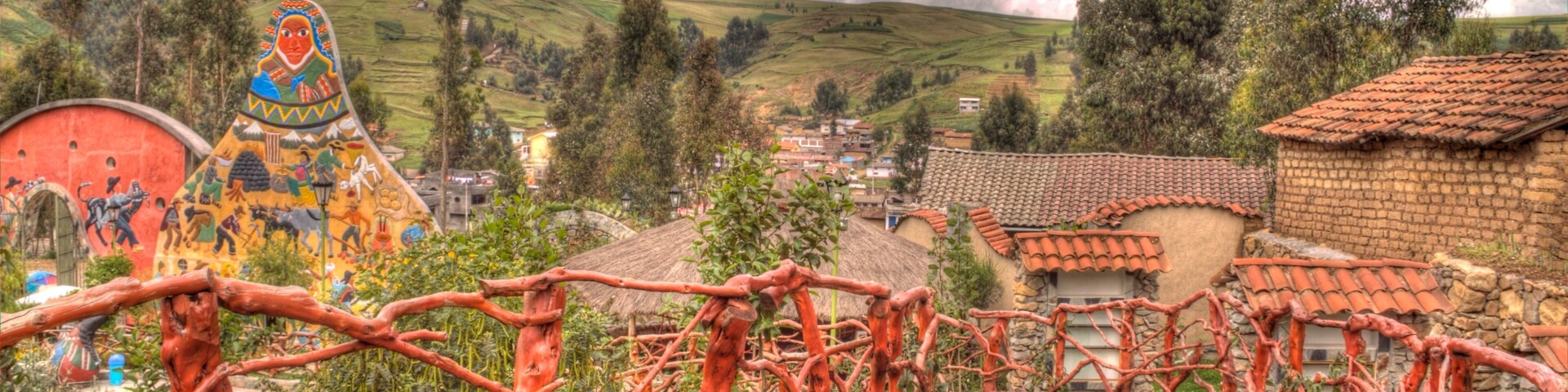 A wooden bridge with red railings, a colorful mural, and a village with terracotta roofs in the background.