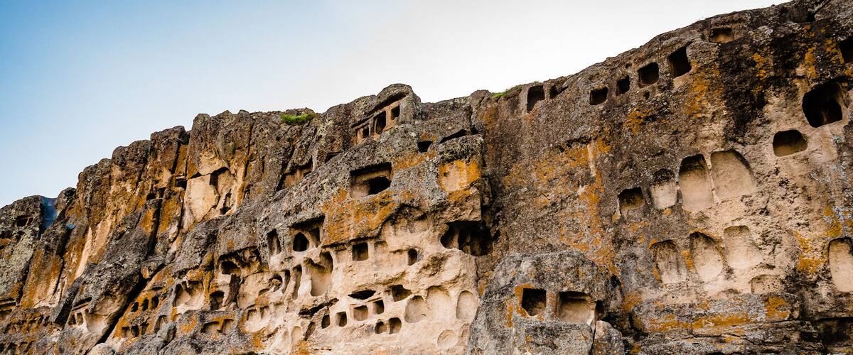 A rock face with numerous cave-like openings and a grassy foreground with cacti.