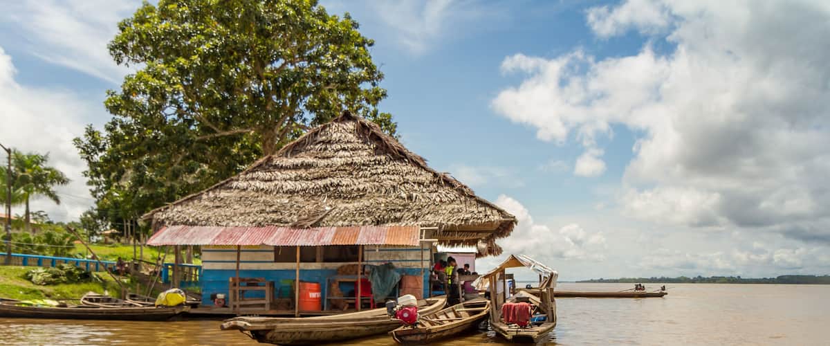 A thatched-roof hut by a calm river with several boats docked nearby.