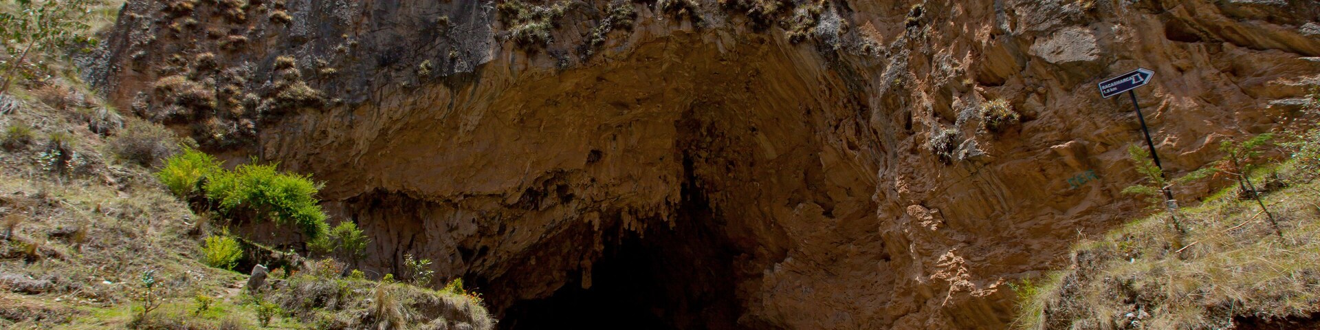 La entrada de una cueva con un sendero de tierra que se adentra, rodeada de terreno rocoso y vegetación escasa.