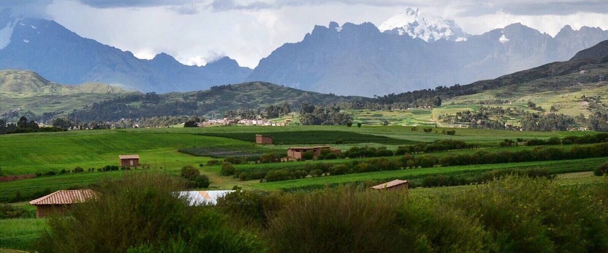 A rural landscape with green fields, scattered farm buildings, and snow-capped mountains in the background.