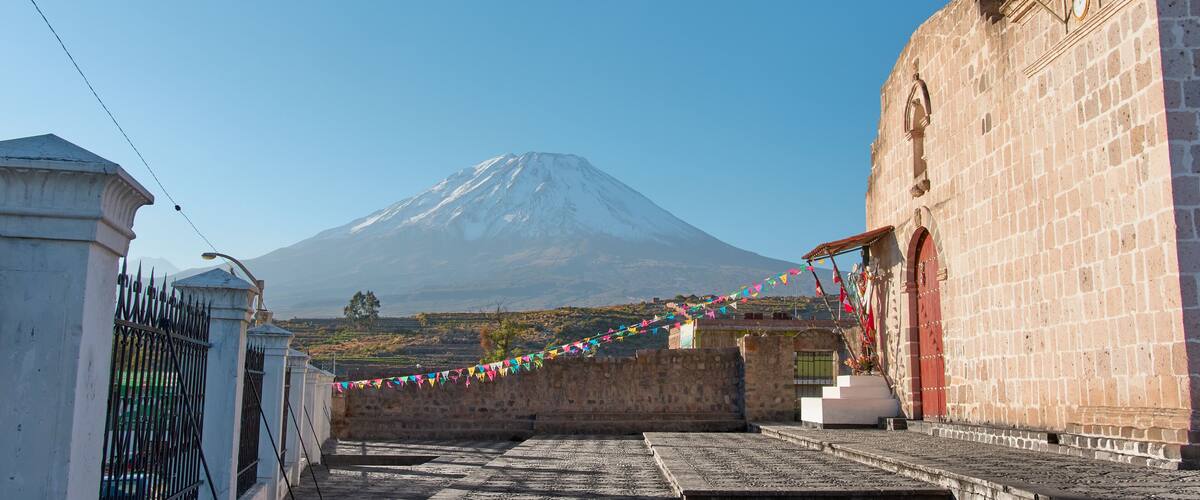A church with a bell tower, a stone wall, and a view of a snow-capped mountain.
