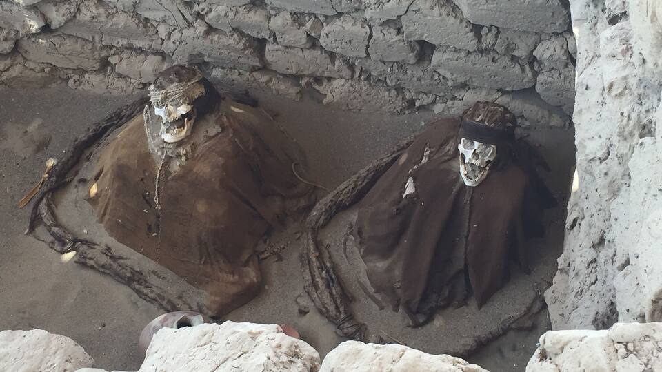 Two mummies in a tomb with stone walls and a sandy floor.